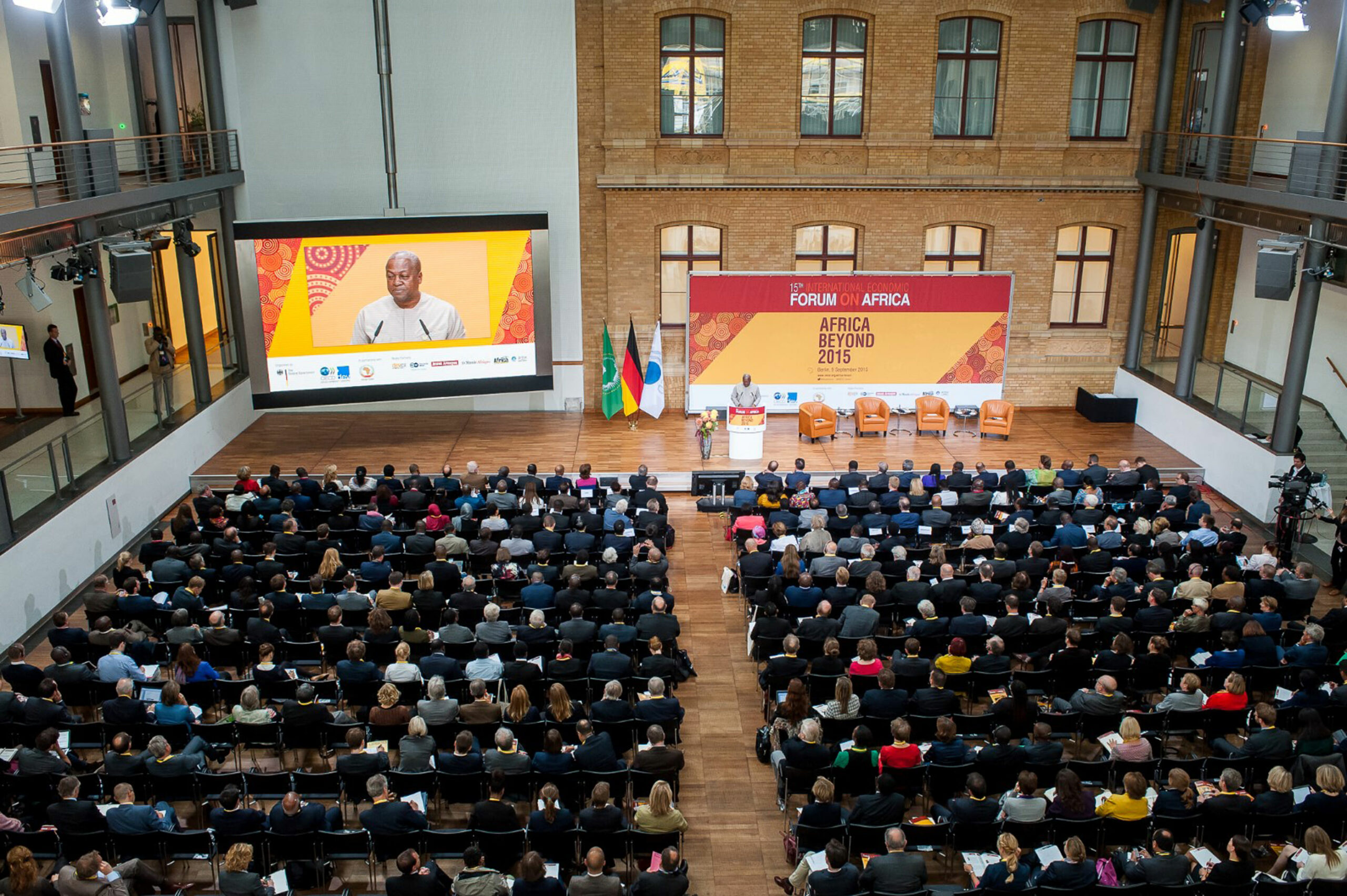 Ein großer Saal mit Publikum, Bühne und großem Bildschirm. Der Redner ist darauf zu sehen